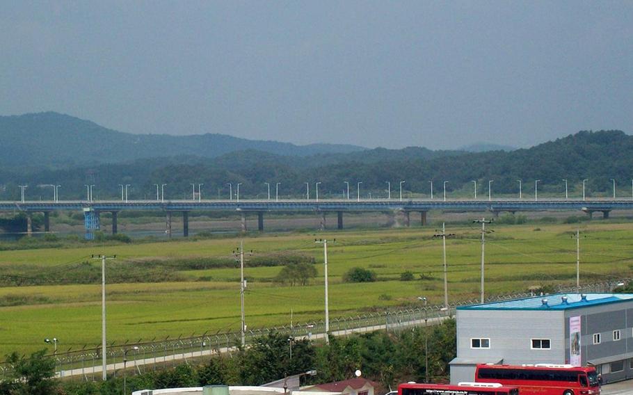 in the foreground, buses are parked, as a long bridge stretches in front of green hills in the background.