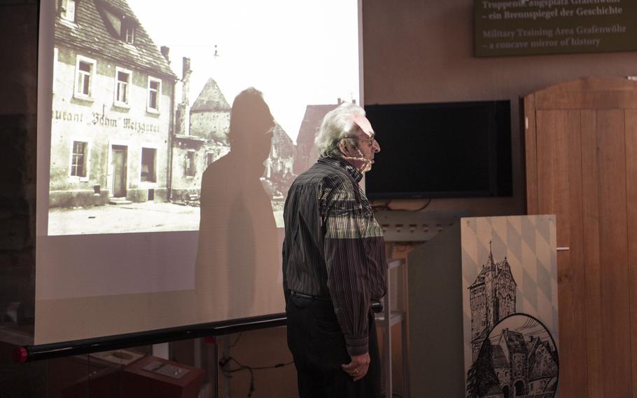 An old man stands in front of a projector screen.