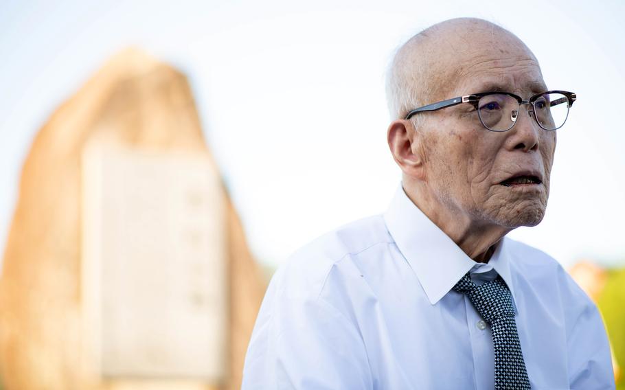 An elderly Japanese man wearing a white shirt, green tie and dark-rimmed glasses stands in front of a blurred stone memorial in the background.