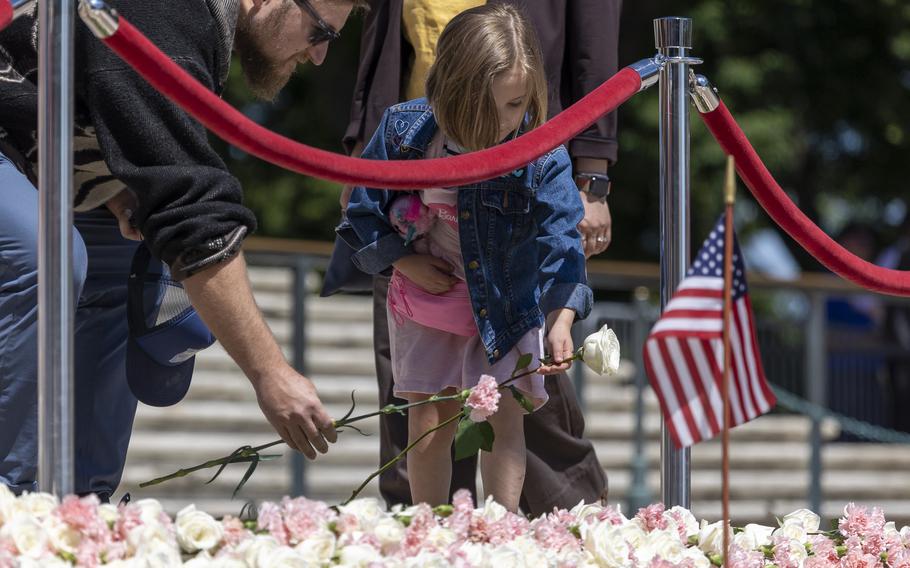 A young girl lays a white flower on the pile of flowers. A man next to her lays a pink flower.