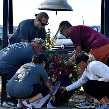 Five people bend over to lay a wreath below a bell.