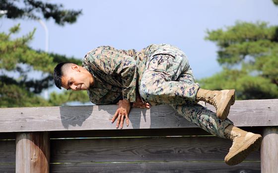 A U.S. service member in camouflage uniform and tan boots climbs over a wooden obstacle during a physical challenge, with trees and a clear sky in the background.