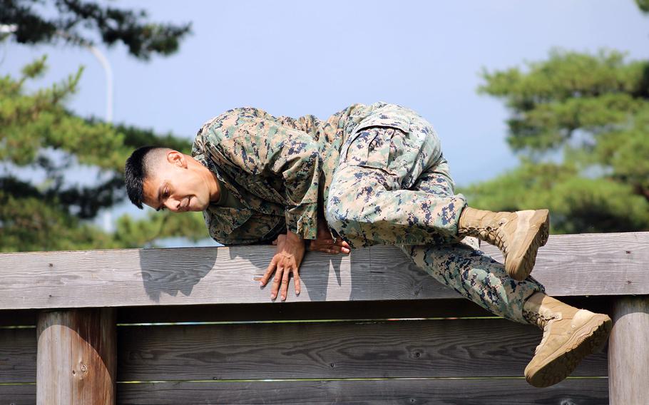 A U.S. service member in camouflage uniform and tan boots climbs over a wooden obstacle during a physical challenge, with trees and a clear sky in the background.