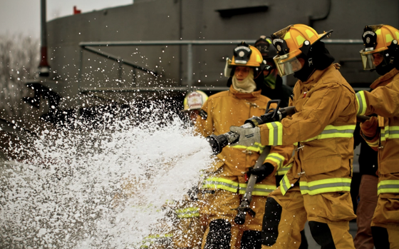 Firefighters in protective suits and helmets spray firefighting foam from a hose.