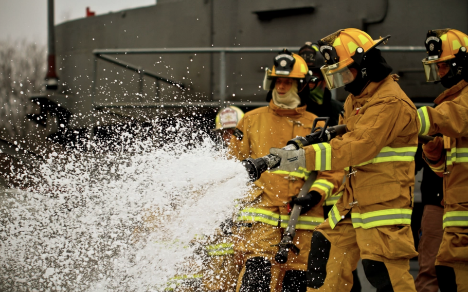 Firefighters in protective suits and helmets spray firefighting foam from a hose.