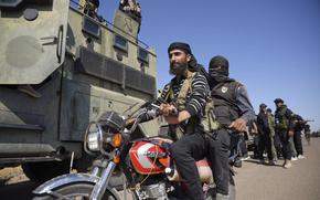 Two men ride a motorcycle next to an armored vehicle.