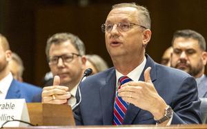 Doug Collins in a suit and red and blue tie, gesturing with both hands while seated and speaking before a microphone.