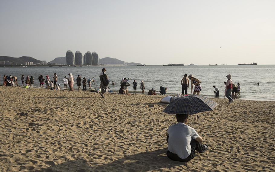 A man sits under an umbrella on Sanya Bay beach as buildings stand in the distance on Phoenix Island, an artificial archipelago developed by Sanya Phoenix Island Development Co., in Sanya, China, on March 13, 2018.