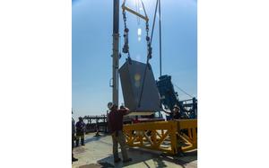 Shipyard workers manage the powered lifting of a large box.