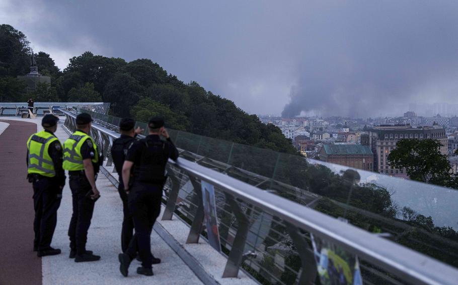 Smoke is seen in the distance rising over a city skyline as uniformed police officers watch from a bridge in the foreground.