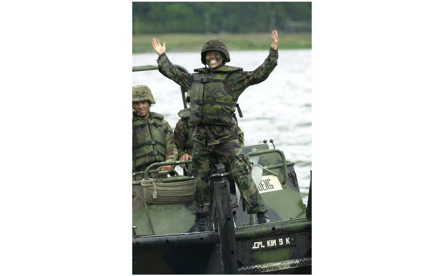 A female soldier grins and stands with her hands raised on a vehicle.