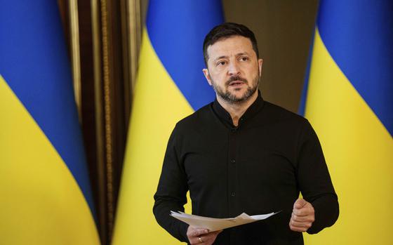 Volodymyr Zelenskyy speaks while gesturing and holding papers in one hand, in front of Ukrainian flags.