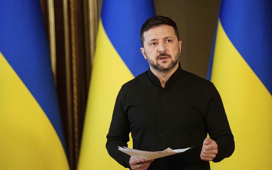 Volodymyr Zelenskyy speaks while gesturing and holding papers in one hand, in front of Ukrainian flags.