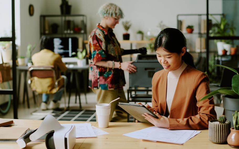 Young Asian manager sitting at desk using digital tablet at work in modern office, her colleagues printing documents and coding in background