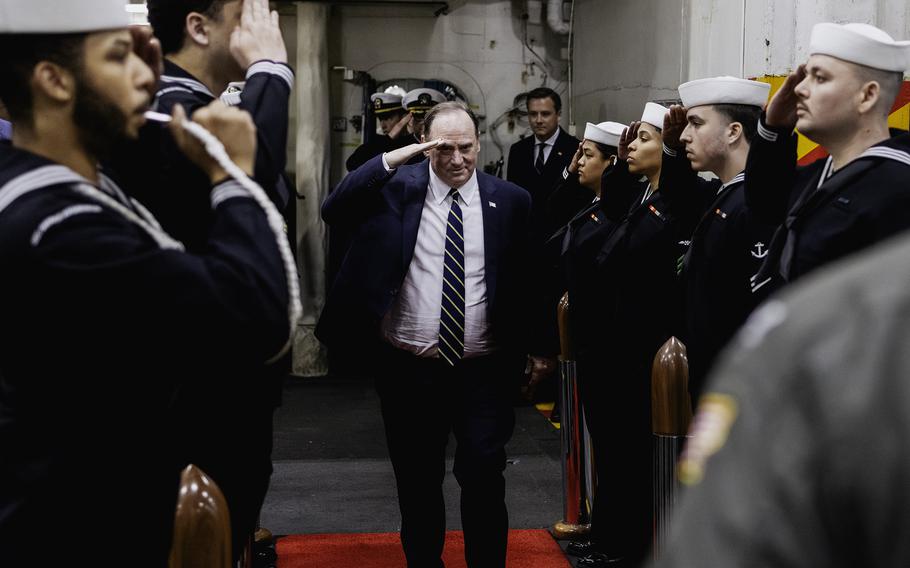 A man in a suit salutes while standing on a red carpet as sailors stand at attention on either side of him and salute.