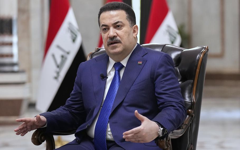 A man in a suit speaks while sitting in a nice chair and gesturing slightly with both hands.