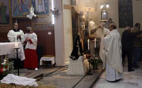 A priest in white robes praying before a crucifix statue.