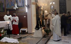 A priest in white robes praying before a crucifix statue.