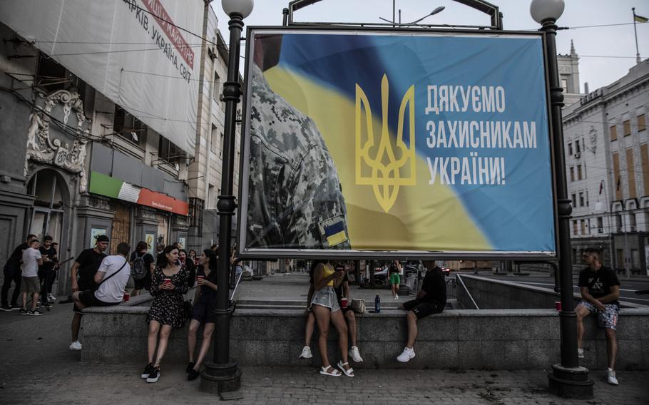 Young Ukrainians drinking outside the Piana Vyshnia, or Drunk Cherry, a Ukrainian chain specializing in sweet cherry liquor near by a giant billboard that reads " Thank you defenders of Ukraine" in the early evening in the city of Kharkiv, Ukraine, on Aug. 11, 2022.