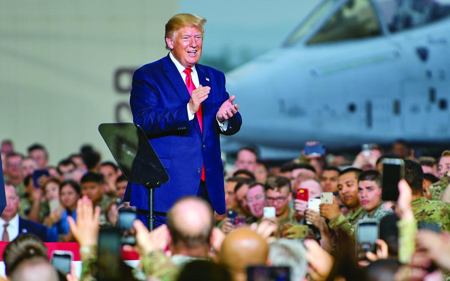 Donald Trump stands on a platform and claps while surrounding by an audience with a military fighter jet in the background.