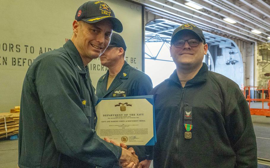 Two Navy officers in blue uniforms and hats shake hands and pose while holding a commendation award between them.