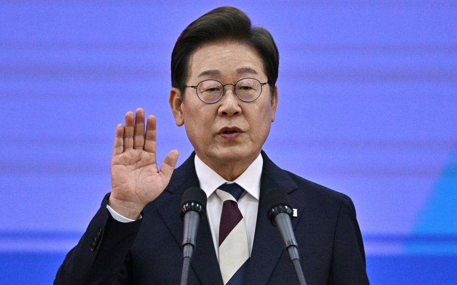 South Korean President Lee Jae-myung, wearing a dark suit and glasses, raises his right hand and speaks into two microphones as he takes the oath of office.