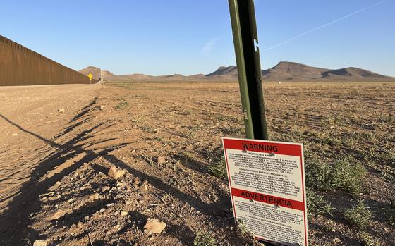 A warning sign mounted on a green metal post stands in an arid desert landscape with brown, rocky terrain.