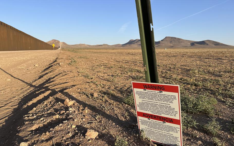 A warning sign mounted on a green metal post stands in an arid desert landscape with brown, rocky terrain.
