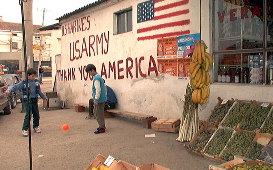Children play soccer next to outdoor produce stands and in front of a wall with “Thank you America” painted on it.