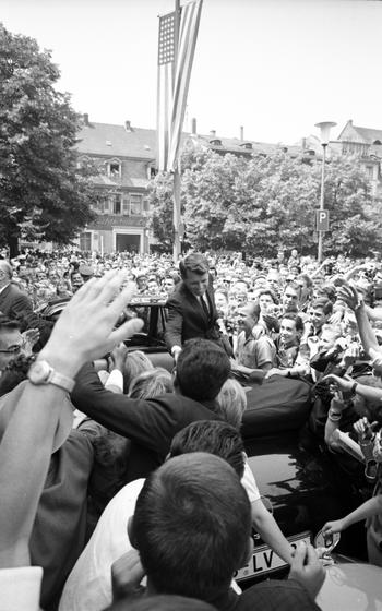 A vertical photo of RFK shaking hands with someone from his car, which is surrounded by fans.