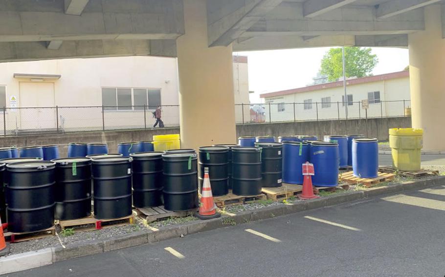 Black and blue barrels sit on a patch of gravel under a concrete overpass with a chain-link fence in the background.