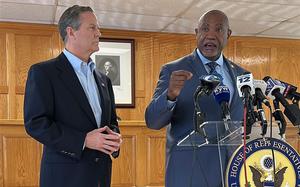 Two men in suits stand in front of a podium with numerous microphones, one of them speaking and the other looking on.