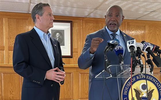 Two men in suits stand in front of a podium with numerous microphones, one of them speaking and the other looking on.