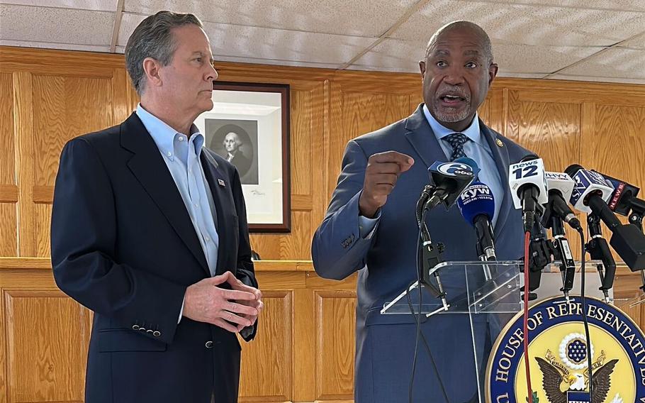Two men in suits stand in front of a podium with numerous microphones, one of them speaking and the other looking on.