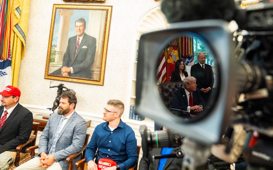 An out-of-focus camera in the foreground with three seated men in-focus farther away.