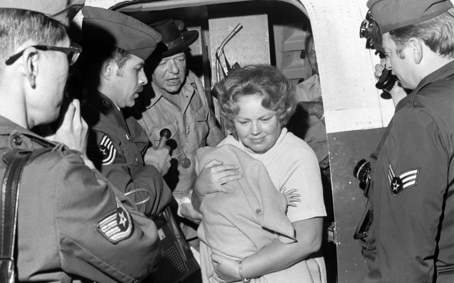 A woman walks out of a plane with a child swaddled in a blanket while men look on.