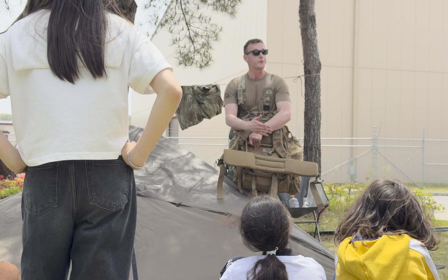 An airman wearing tactical gear and sunglasses speaks to a group of young students seated in front of him. The scene takes place outdoors, next to a tent setup with military gear hanging nearby. 