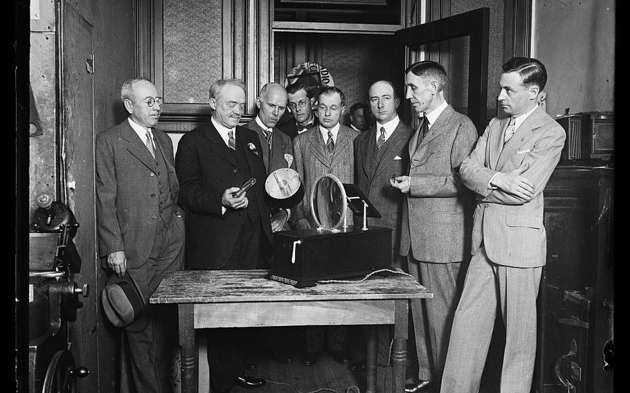 A black and white photo of men in suits standing at a table with a device on it.