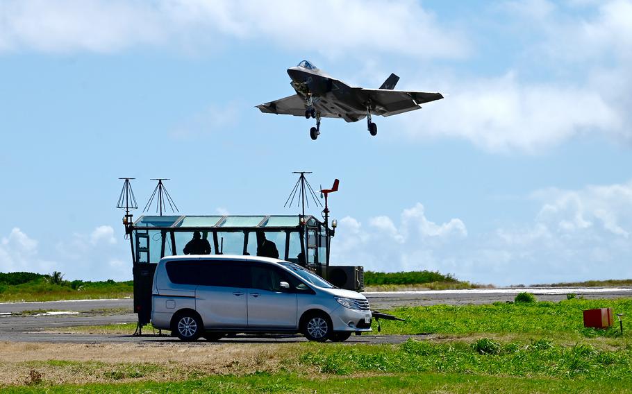 A military fighter jet descends toward a runway with an observation hut and van next to the runway below the jet.