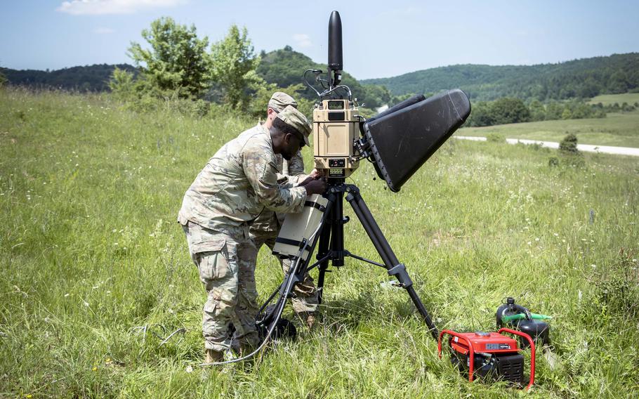 Two soldiers set up a device on a tripod in a grassy area.