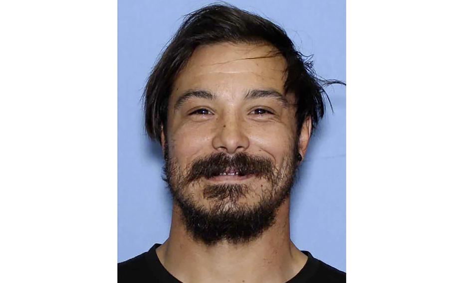 A police booking photo shows a man with dark hair and beard, seen from the neck up, in front of a blue background.