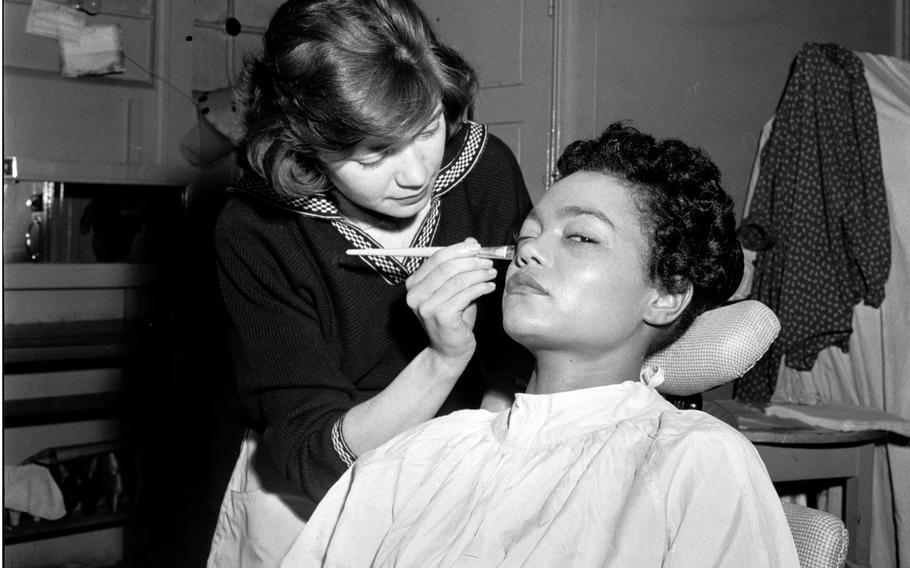 A black-and-white, 1950s-era image of a woman sitting in a dressing room with her head tilted back as a makeup artist uses a brush to apply makeup to her cheek.