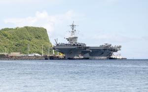 An aircraft carrier docks at a pier with a tree-covered ridge to the left and blue sky and the horizon in the background.