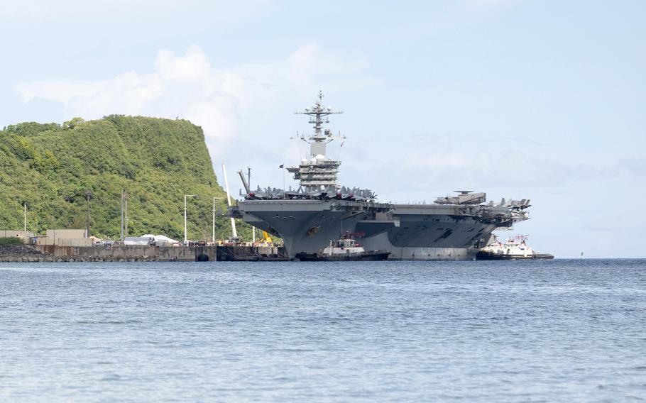 An aircraft carrier docks at a pier with a tree-covered ridge to the left and blue sky and the horizon in the background.