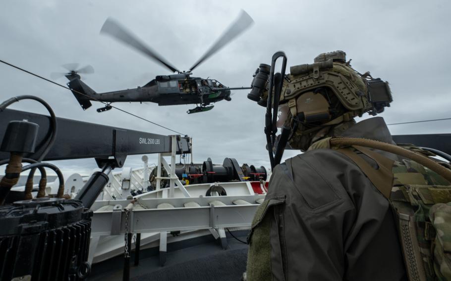 A service member on the deck of a ship looks at something as a helicopter hovers near them.