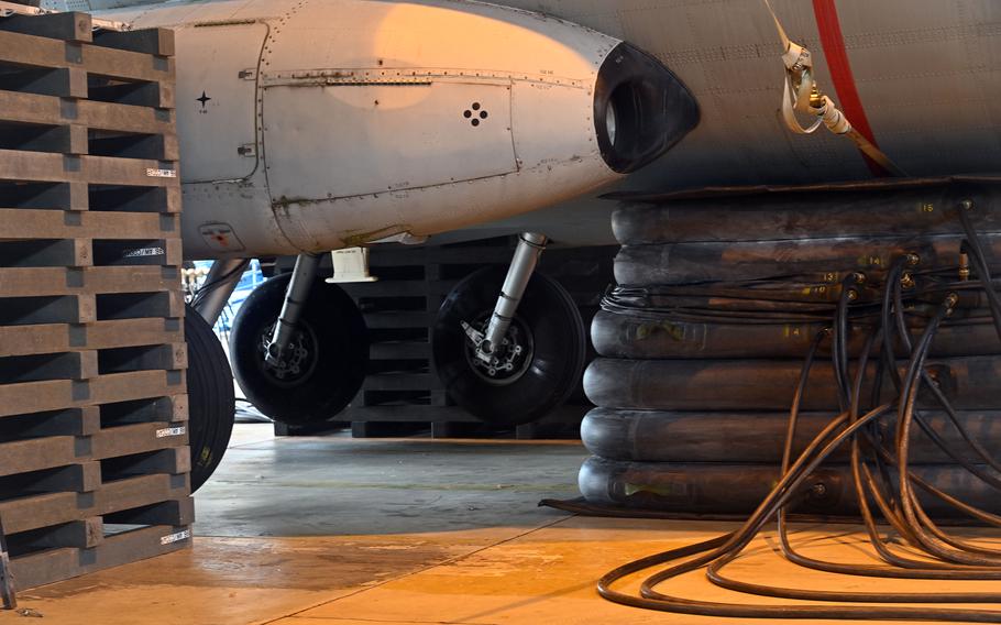 Close-up view underneath a military aircraft showing the nose landing gear and front fuselage. The aircraft is supported by multiple black inflatable air cushions arranged in rows. Orange cables and testing equipment are visible on the hangar floor, with wooden crating materials stacked to the left side.