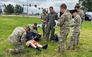 Two military service members in camouflage uniforms kneel next to a patient with blood on his shorts as five other service members stand next to them and observe.