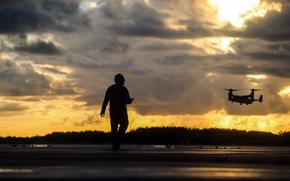 A silhouetted figure in military gear stands on a hill against a dramatic golden sunset sky filled with layered clouds. In the distance, a  Osprey tiltrotor aircraft flies across the horizon. 