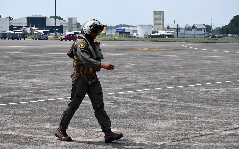 A pilot in flight gear walks across the runway after exiting a fighter jet, with several aircraft and airport facilities visible in the background.