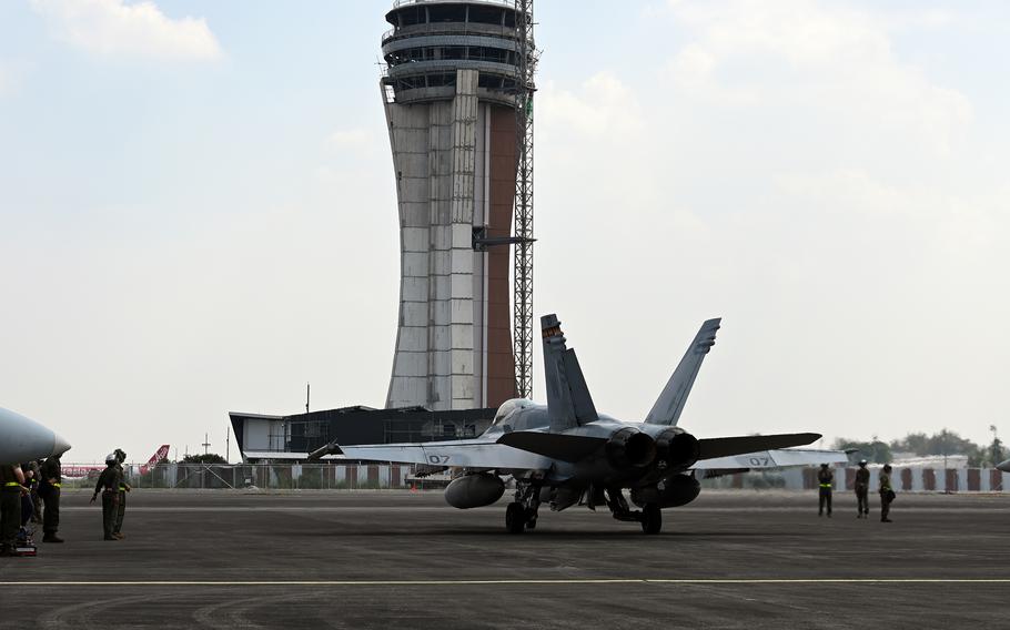 A F/A-18 Hornet jet taxis on a runway in front of an air traffic control tower with military personnel standing nearby on the tarmac.